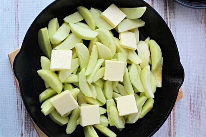 Sliced apples topped with butter pats in a black skillet on a light wooden surface, solid for preparing Pork Chops with Apples or your favorite Baked Pork Chops Recipe.
