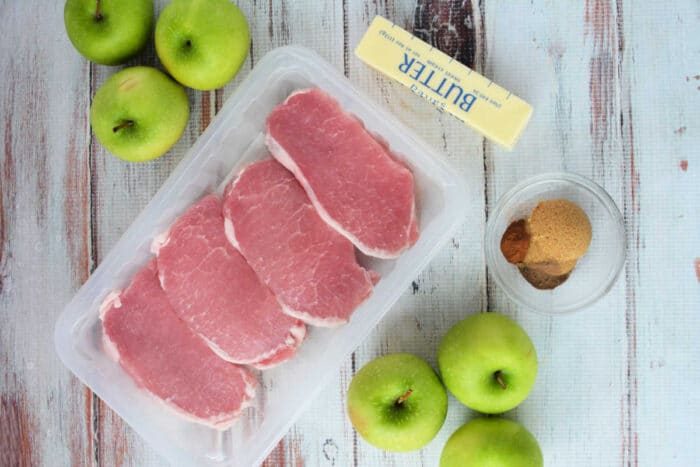Raw pork chops in a plastic tray, green apples, a stick of butter, and brown sugar with spices on a table—solid for trying a good baked pork chops recipe.