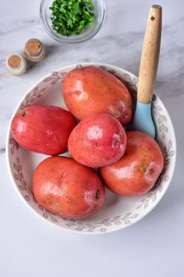 A white bowl with a floral pattern contains five red potatoes and a light blue vegetable peeler with a wooden handle, solid for prepping baked red potatoes. In the background, on a marble surface, there's a small bowl of chopped herbs and two spice jars.