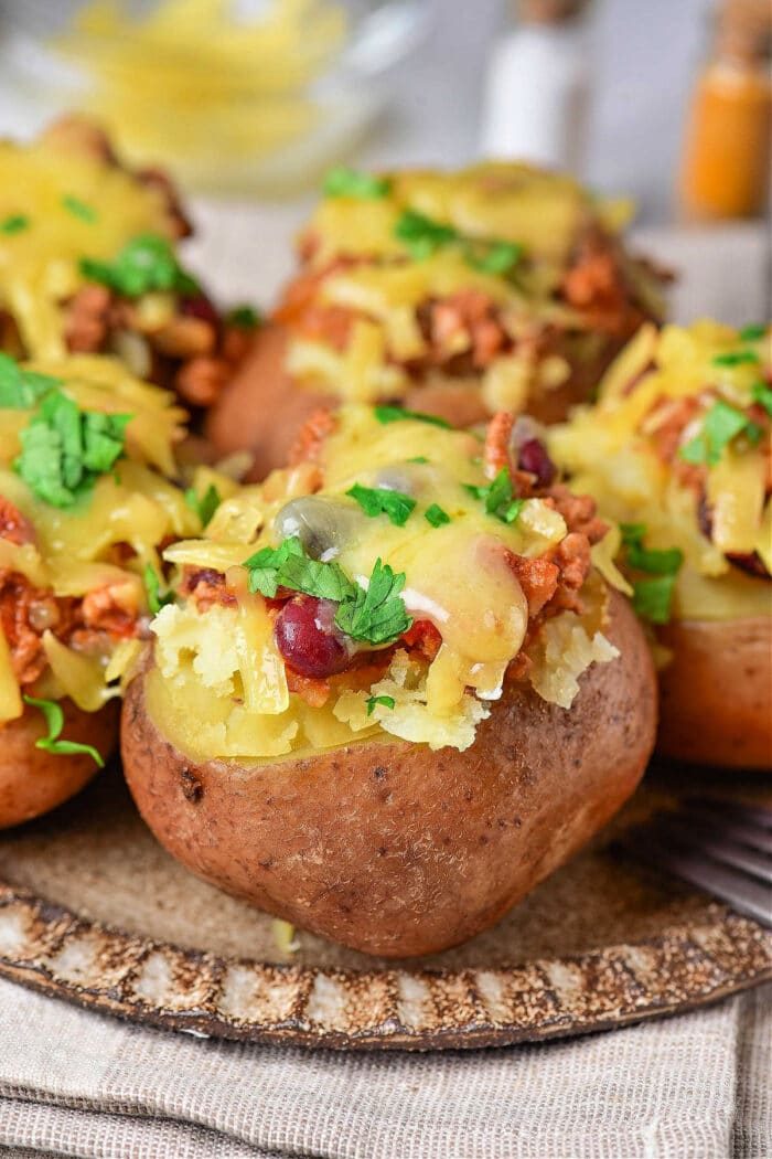 A close-up of baked red potatoes topped with chili, melted cheese, and garnished with chopped cilantro on a plate. The potatoes are arranged on a beige cloth, with a blurred background featuring a bowl and condiment containers.