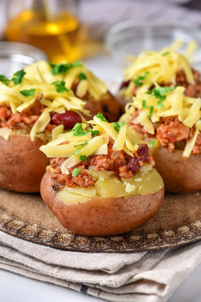 Three baked red potatoes topped with chili, shredded cheese, and chopped parsley are displayed on a rustic, round wooden plate. The plate is placed on a folded beige cloth. In the background are glass containers, slightly out of focus.