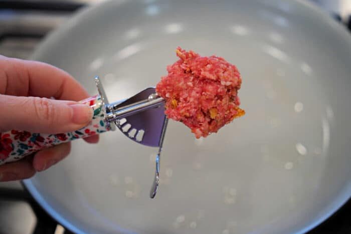 Hand holding a scoop of raw ground meat over an empty frying pan on a stove, solid for preparing baked spaghetti with meatballs.