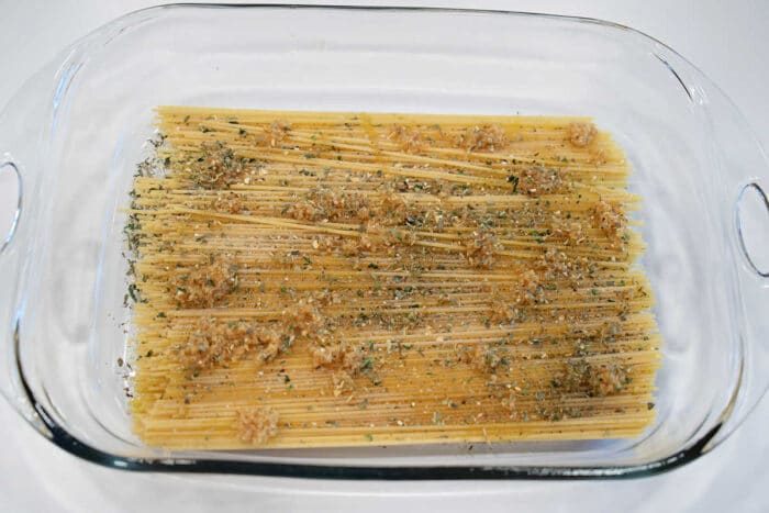 Uncooked spaghetti in a glass baking dish, topped with minced garlic and dried herbs, ready to be transformed into a classic baked spaghetti with meatballs.