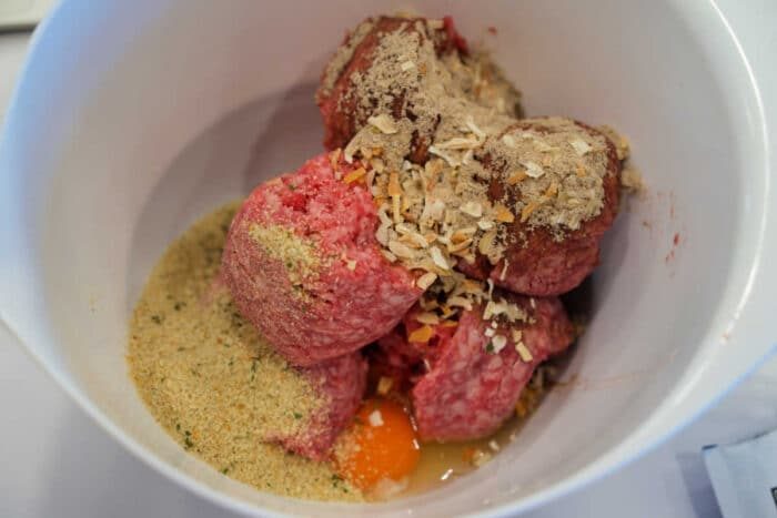 Raw ground meat, breadcrumbs, spices, and a cracked egg in a white mixing bowl, ready to be mixed for good meatballs—solid for pairing with baked spaghetti.