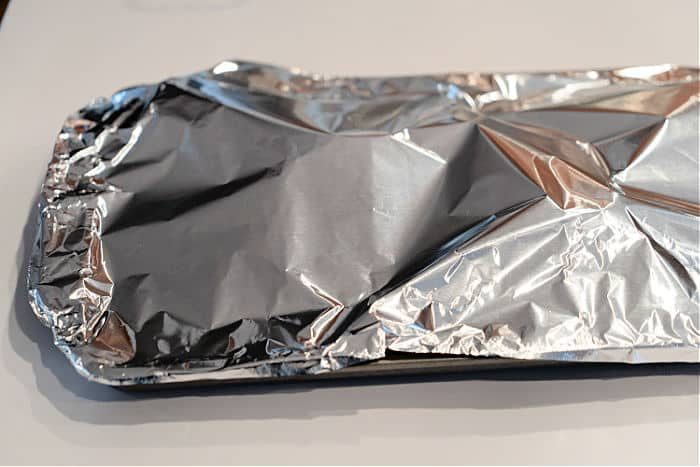 A rectangular baking pan covered tightly with shiny aluminum foil, solid for preparing good baked turkey legs, sits on a white surface.
