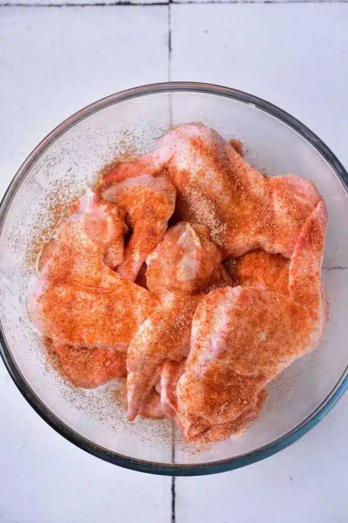 A glass bowl containing raw chicken wings, seasoned with a reddish spice mix, ready to be transformed into good baked whole chicken wings, sits on a white tiled surface.