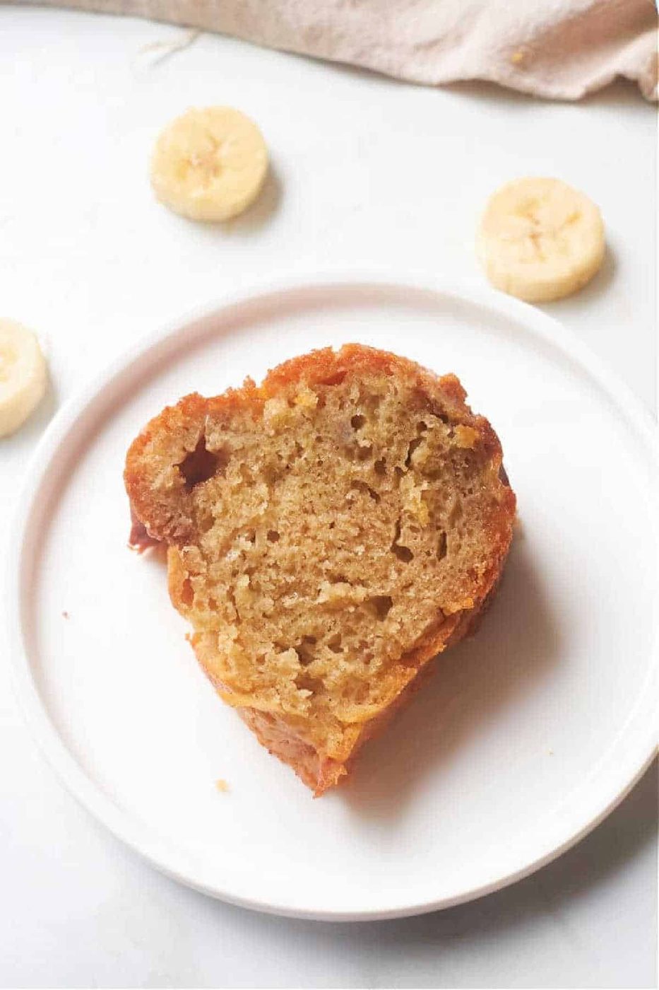 A slice of Banana Bread Bundt Cake sits on a white plate with banana slices in the background.