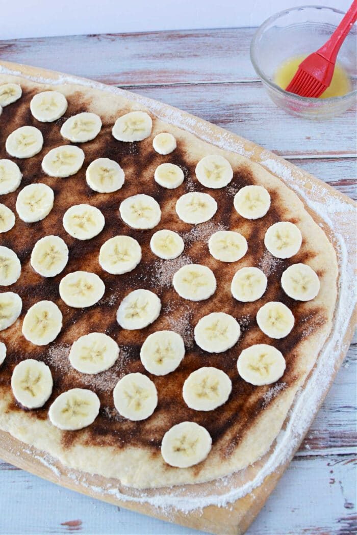 A wooden surface cradles cinnamon roll dough topped with a blend reminiscent of banana bread cinnamon rolls, featuring cinnamon, sugar, and sliced bananas.