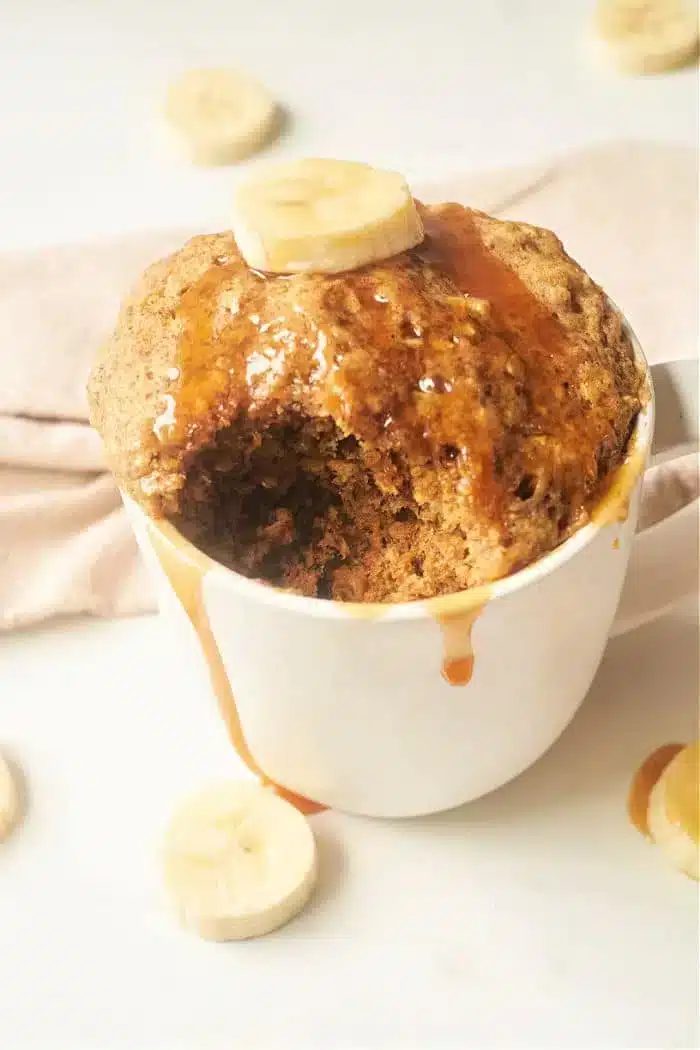 A Banana Bread in a Mug creation, topped with a banana slice and drizzled with syrup. The cake has a bite taken out of it, revealing its moist texture. Additional banana slices are artfully scattered around the mug on a light surface, enhancing its good appeal.