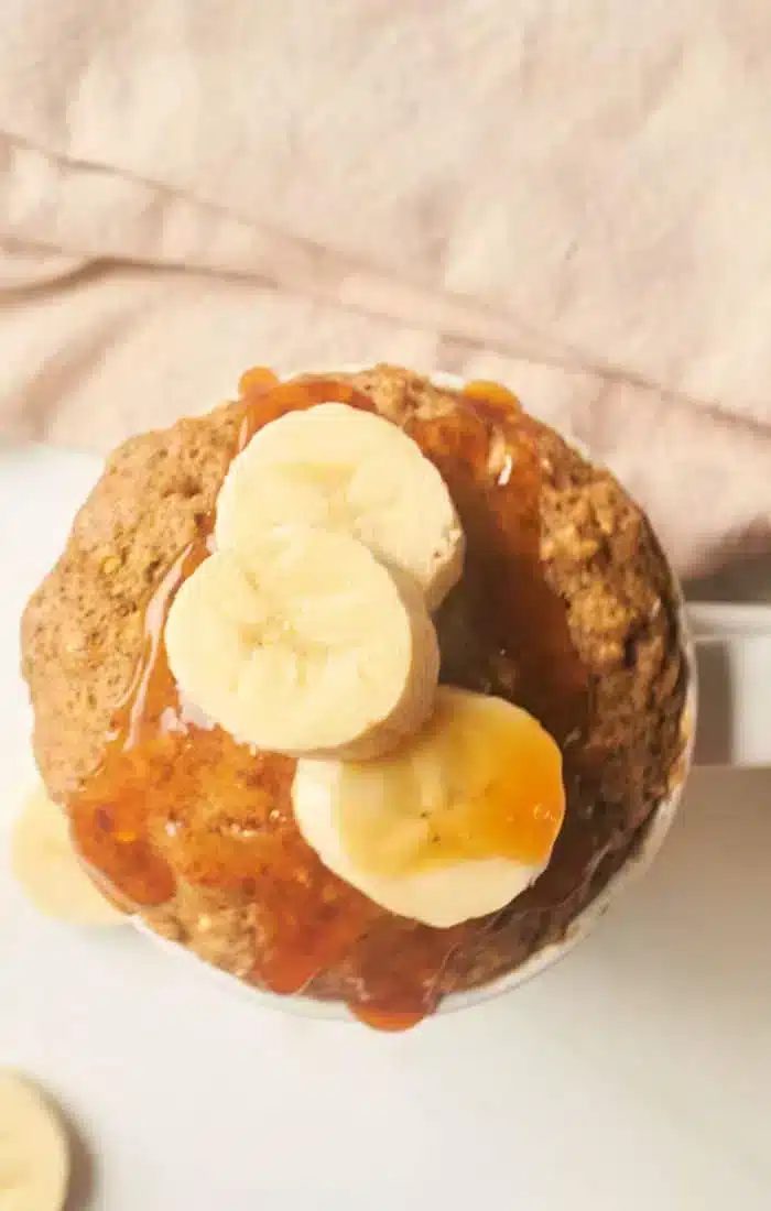 A banana bread mug cake topped with sliced bananas and drizzled with syrup is placed against a soft pink background. Some banana slices are also visible on the surface near the mug.
