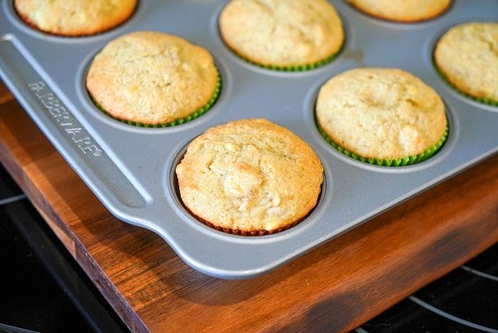 A tray of freshly baked banana bread muffins, made with a surprising hint of mayonnaise, rests in a metal muffin tin on a wooden surface.