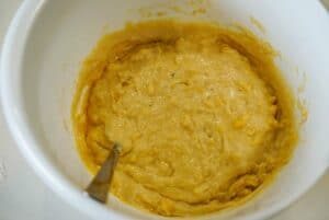 Thick yellow batter for banana bread with mayonnaise being mixed in a white bowl with a metal spoon.