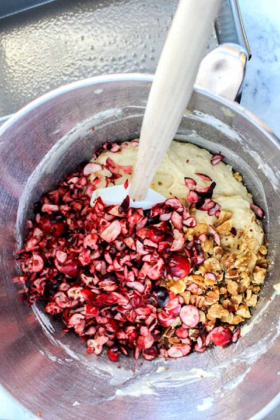 Chopped cranberries and walnuts are being mixed into batter for good banana cranberry bread in a metal bowl with a spatula.