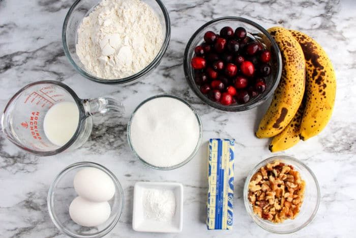 A medley of baking ingredients spread across a marble surface: ripe bananas, cranberries, flour, sugar, eggs, crunchy walnuts, creamy butter, milk, and baking powder—all coming together to inspire banana cranberry bread.