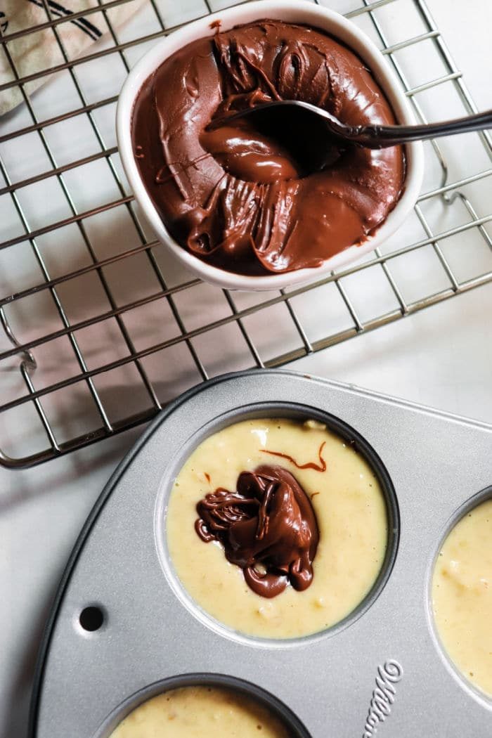 Muffin batter in a pan with a dollop of chocolate, next to a bowl of melted chocolate on a cooling rack, transforms into delightful banana Nutella muffins solid for any indulgent moment.