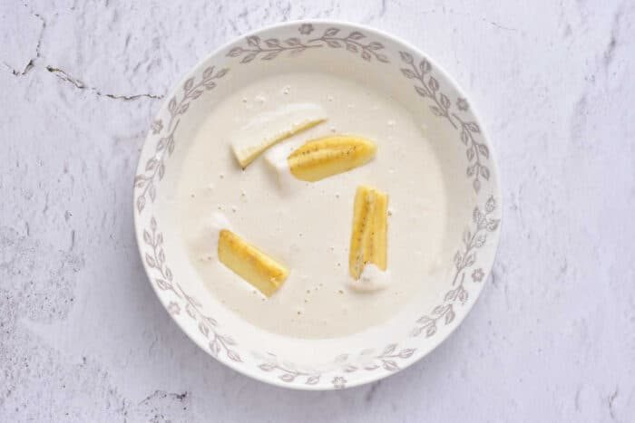 A bowl of tempura is adorned with banana slices, reminiscent of delicate banana tempura, set against a white, textured background.