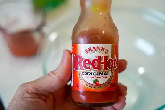 A hand holds a bottle of Frank's RedHot Original Cayenne Pepper Sauce with its signature red label. In the blurry background, a measuring cup and glass bowl hint at a good creation—perhaps the solid bang bang shrimp sauce in the making.