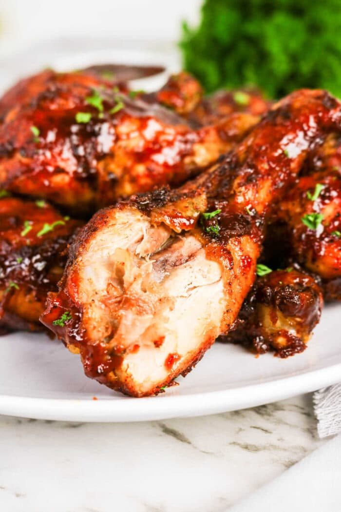 Oven baked BBQ chicken legs are served on a white plate, one with a bite taken out, and garnished with chopped herbs—a good addition to your collection of chicken recipes.