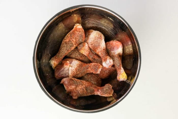 Raw chicken drumsticks coated with spices in a metal mixing bowl, viewed from above on a white background—solid for preparing oven baked chicken or BBQ chicken legs.