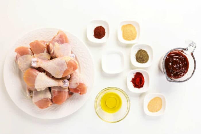 Raw chicken drumsticks on a plate with small bowls of spices, oil, salt, and barbecue sauce on a white background—solid for your next oven baked BBQ chicken legs recipe.