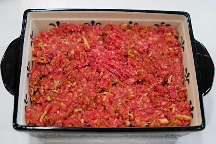 Raw ground beef mixed with onions is spread evenly in a rectangular baking dish, solid for a classic meatloaf recipe and ready for cooking.