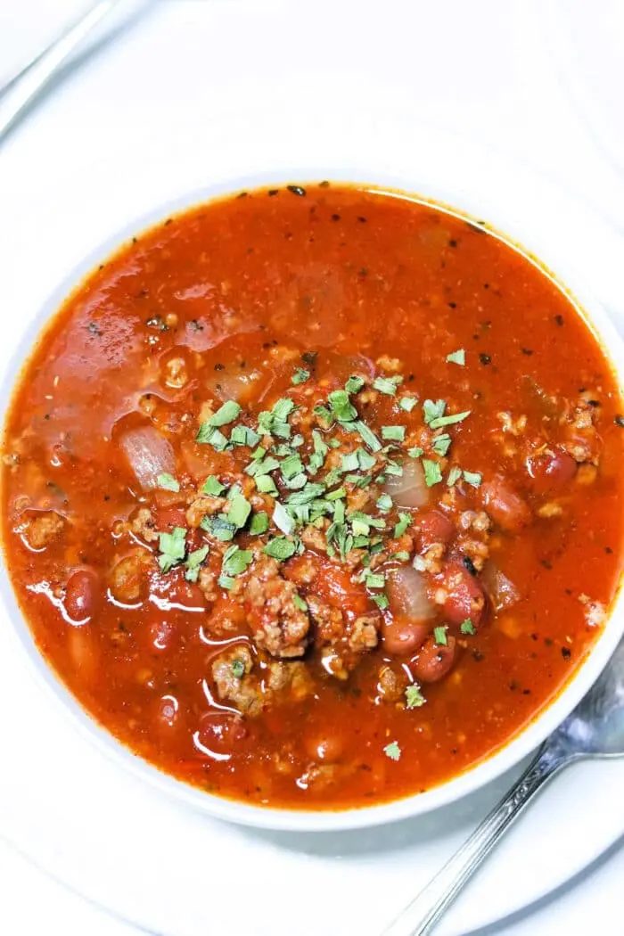 A bowl of chunky bear meat chili topped with chopped herbs. The hearty mix features beans, onions, and tender ground meat, all served in a pristine white bowl with a spoon ready on the side.