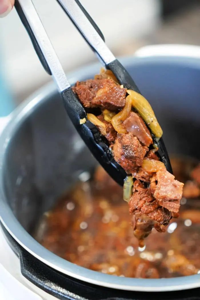 Tongs holding cooked pieces of beef and onions over a pot filled with broth suggest a good beef arm roast recipe. The meat appears tender, perfectly mixed with onions, indicating it's been lovingly simmering in rich liquid.