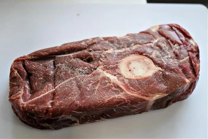 A raw cut of beef arm roast on a white surface, featuring a bone in the center. The meat has a marbled texture and a reddish-brown color, solid for your next savory recipe.