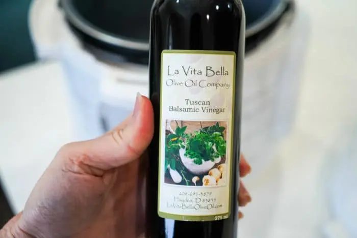 A hand holds a bottle of La Vita Bella Olive Oil Company's Tuscan Balsamic Vinegar, solid for enhancing your beef arm roast recipe. The label features images of herbs and garlic, beautifully showcased against a blurred background.
