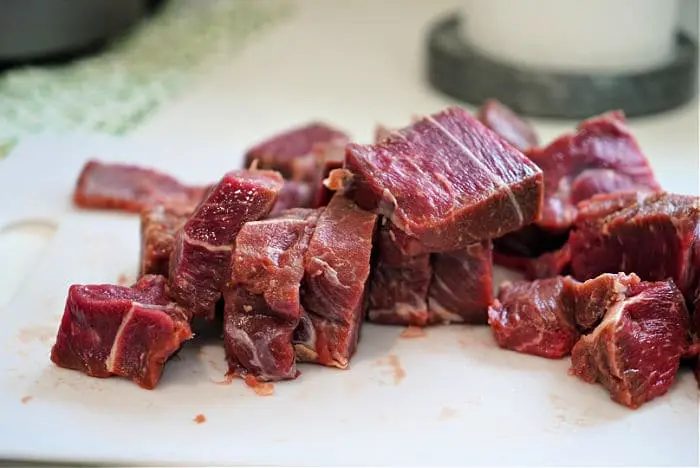 Chopped pieces of raw beef arm roast pieces are placed on a white cutting board, solid for starting a beef arm roast recipe. The meat is marbled with fat, and the board rests on a kitchen counter. The background features a green-patterned cloth and part of a round, gray object.