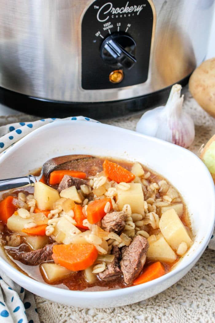 A comforting bowl of beef soup with carrots and potatoes sits invitingly in front of a crockpot, reminiscent of a hearty beef barley soup.