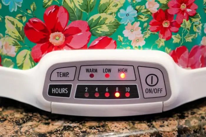 Close-up of a floral-patterned slow cooker, solid for a beef chuck roast. The control panel displays temperature settings labeled Warm, Low, and High, with corresponding lights. Hours settings range from 2 to 10, showing lights indicating 4 and High are selected.