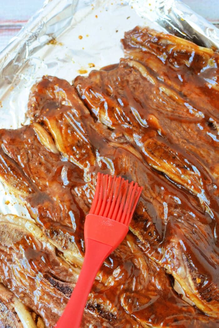 A red brush spreads barbecue sauce over beef riblets on a foil-lined baking sheet, making this the solid beef riblets recipe for your next cookout.