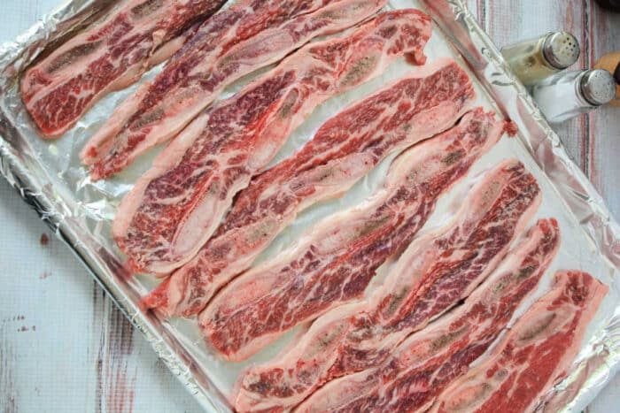 Raw marinated beef riblets arranged on a foil-lined baking sheet, with salt and pepper shakers nearby—solid for trying out your favorite Beef Riblets Recipe.