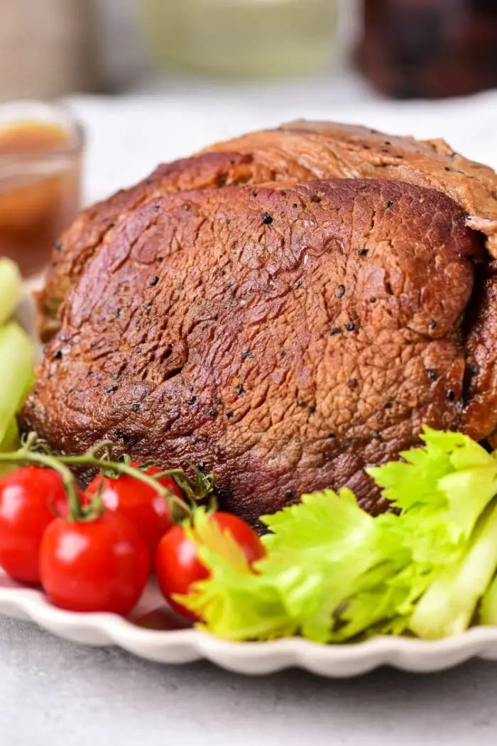 A roasted beef roast shoulder served on a white plate, garnished with fresh cherry tomatoes and celery stalks. The beef appears well-seasoned with a brown crust. A blurred bowl of sauce is visible in the background.
