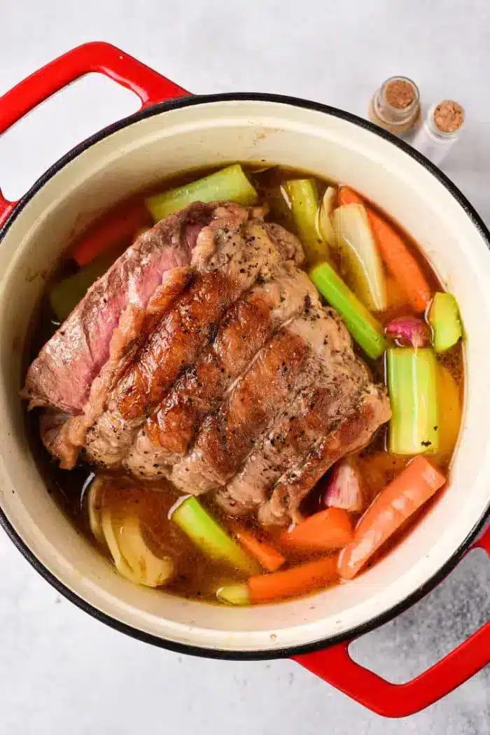 A beef roast is cooking in a red cast iron pot, surrounded by various vegetables including carrots, celery, and onions, all simmering in a rich brown broth. The tender shoulder cut enhances the flavor perfectly. Two small spice containers are visible in the background.