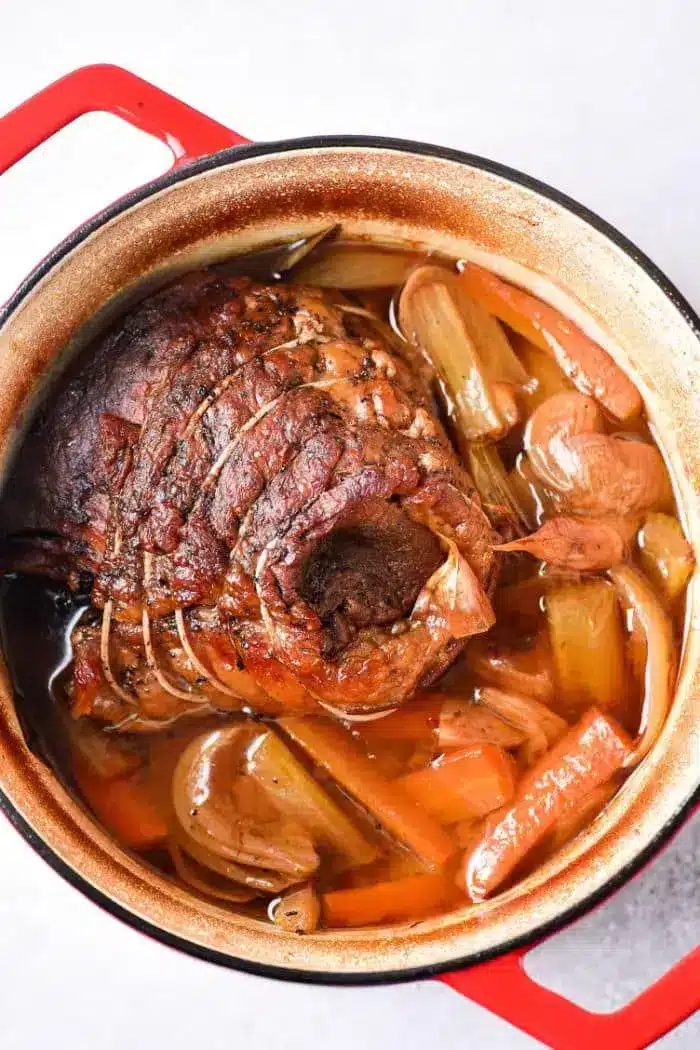 A cooked beef roast shoulder in a red Dutch oven, surrounded by chunks of carrots, celery, and onions in a brown broth. The meat is seasoned and tied with string, resting in a savory liquid with visible spices.