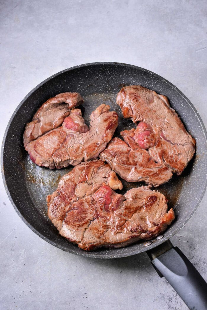 Three pieces of seared steak, solid for a Beef Shin recipe, cooking in a black frying pan.
