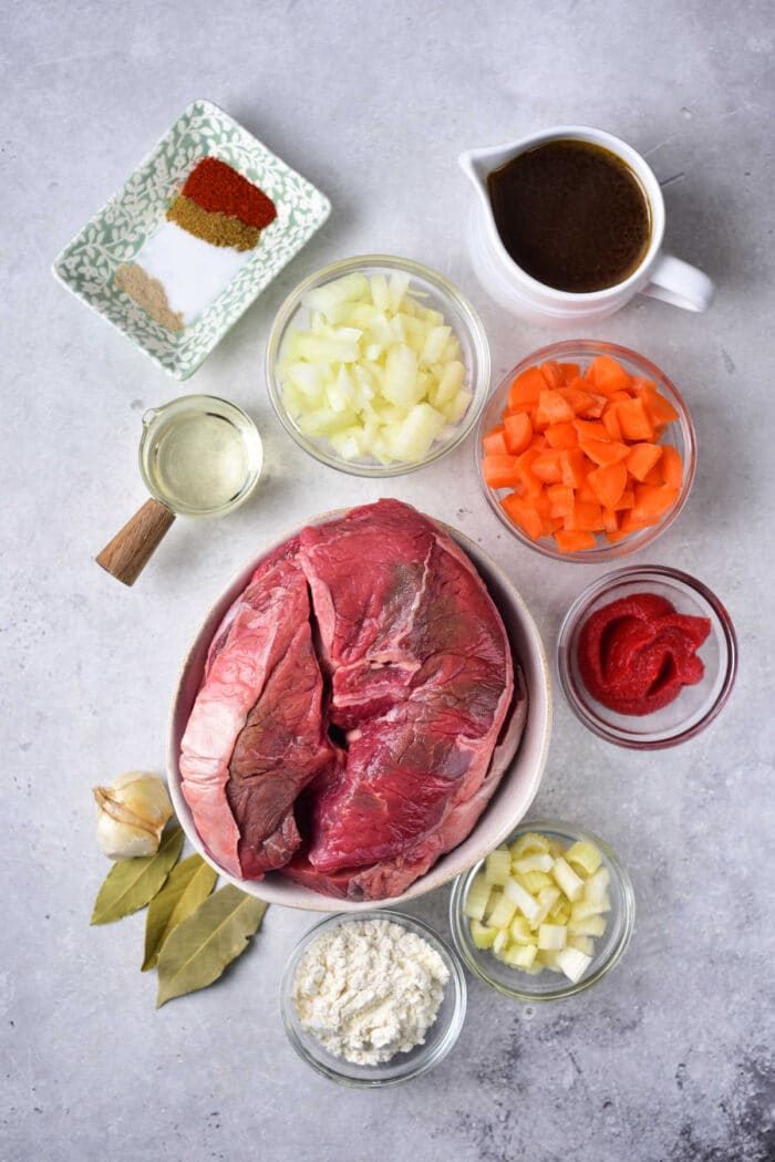 Ingredients for a hearty beef stew: tender beef shin, chopped onions and carrots, spices, broth, tomato paste, cream, oil, garlic, and bay leaves.