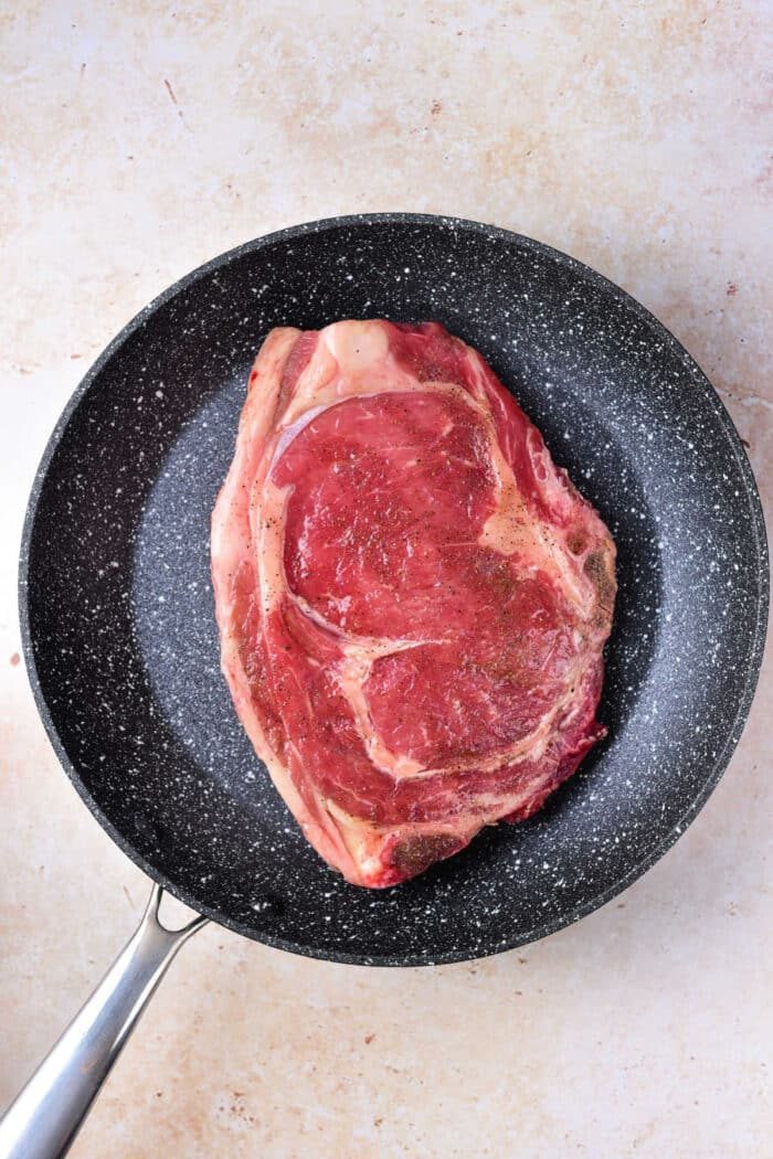 Raw seasoned steak in a black frying pan on a light-colored countertop, illustrating the first step in How to Cook Beef Strip Steak in the Oven.