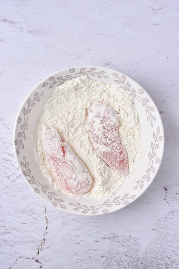 Two raw chicken tenders, are coated in flour and elegantly arranged in a decorative bowl on a light textured surface.