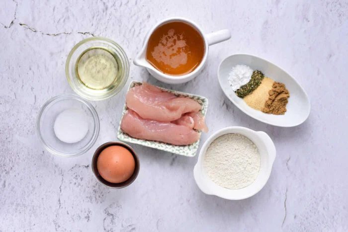 Raw chicken, an egg, flour, seasonings, oil, salt, and broth in bowls on a light textured surface—solid for making crispy beer battered chicken tenders.