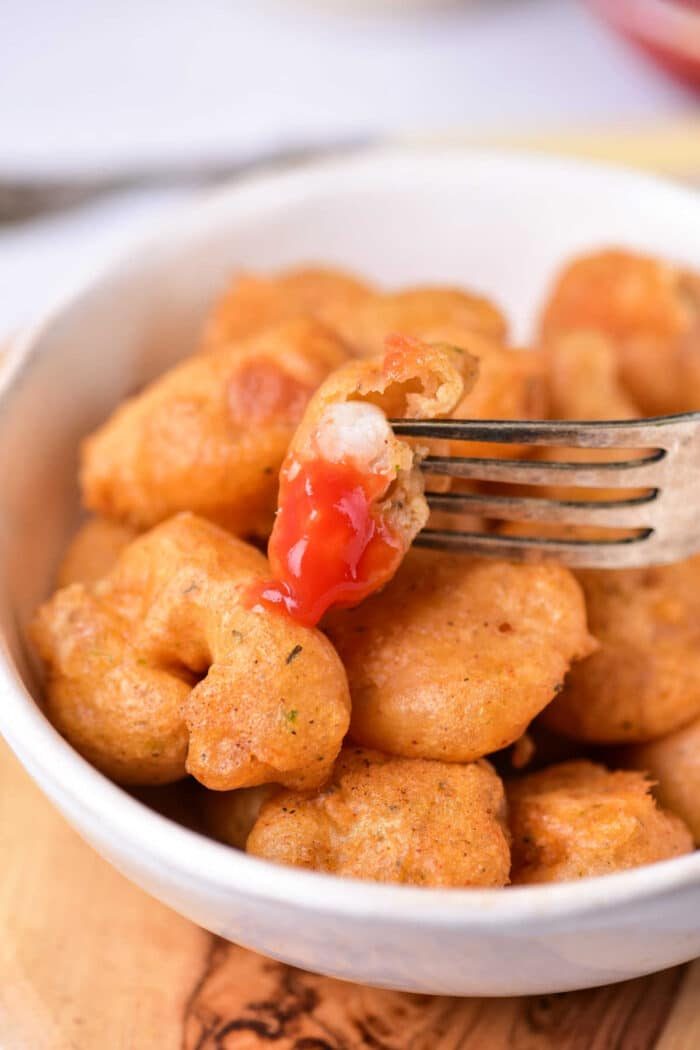A fork holds a beer-battered fried shrimp bite with ketchup in a white bowl of similar bites.