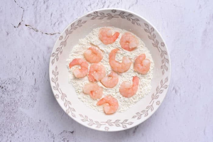 A white bowl with a decorative border contains a mixture of flour and several uncooked shrimp placed on the surface, ready to be transformed into good Beer Batter Shrimp. The bowl sits on a light, textured background.