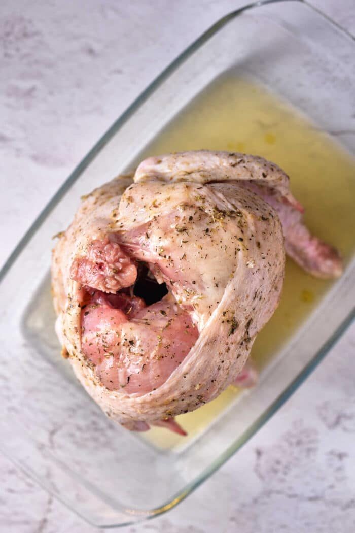 Raw seasoned Beer Can Chicken in the Oven, sits ready in a glass baking dish on a light surface.