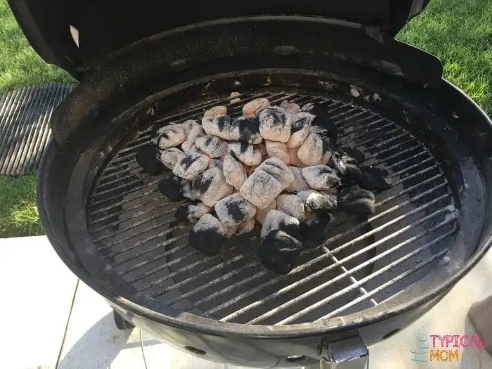A round charcoal grill with its lid open reveals a pile of partially burned charcoal briquettes, solid for preparing beer can chicken. The backdrop features some grass and patio tiles, setting the scene for a relaxed backyard barbecue.