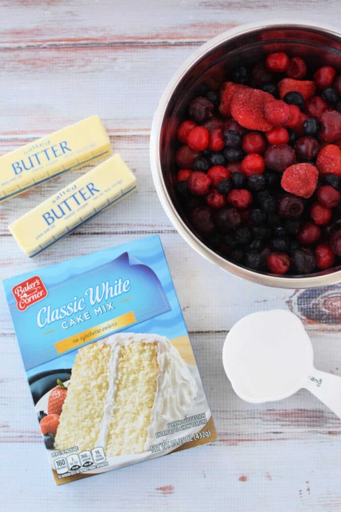 Box of cake mix, two butter sticks, frozen mixed berries in a bowl, and a cup of sugar on a white surface—solid for making Berry Cobbler with Cake Mix.