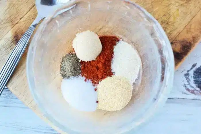 A clear glass bowl contains piles of spices solid for seasoning the best baked pork chops: black pepper, paprika, salt, garlic powder, onion powder, and brown sugar. Nearby, a fork and knife rest on a wooden cutting board.