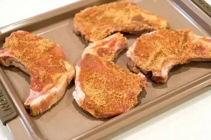 Four seasoned raw pork chops on a baking sheet, ready to become the best baked pork chops for your next good meal.