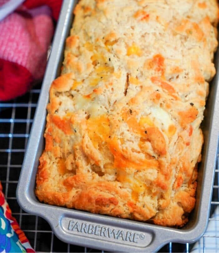 Beer cheddar bread in a metal loaf pan, cooling on a wire rack beside colorful towels, captures the essence of the best beer bread recipe.